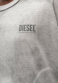 Κοντινό πλάνο ανοιχτογκρι πουκαμίσου με το λογότυπο "DIESEL For Successful Living" τυπωμένο στο στήθος, που φοριέται από ένα άτομο.