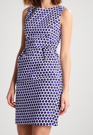Donna che indossa un vestito attillato senza maniche con motivo a pois blu e bianco, in piedi davanti a uno sfondo uniforme.