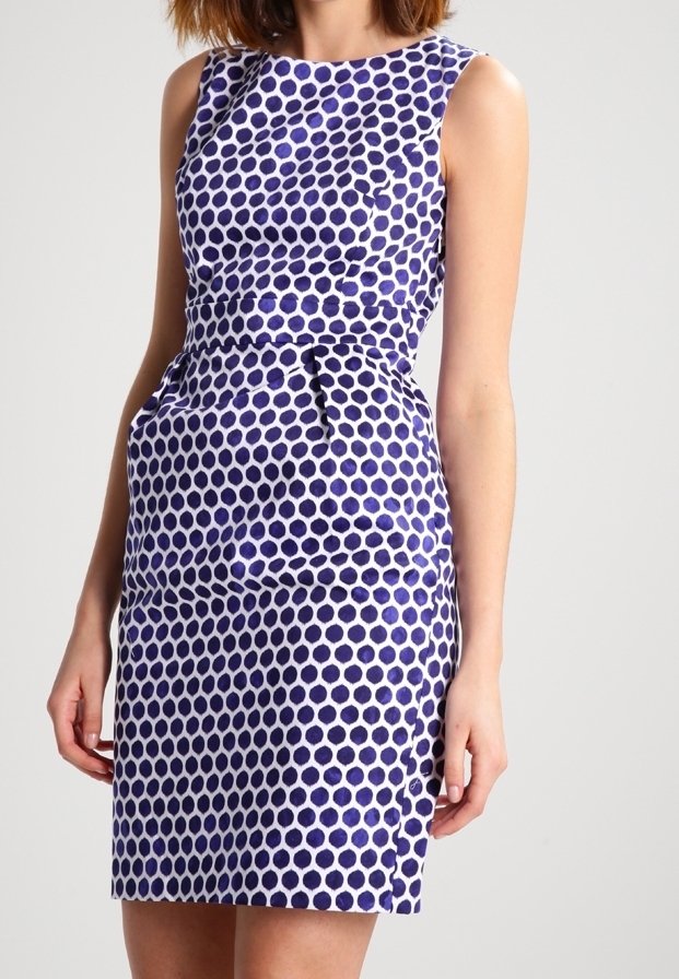 Femme portant une robe sans manches, ajustée, avec un motif à pois bleu et blanc, debout devant un fond uni.