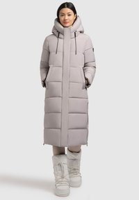 Dolga siva puffer jakna z visokim ovratnikom, sprednjim zadrgo in dvema stranskima žepoma. Ima kapuco in je v kombinaciji z belimi izoliranimi škornji.