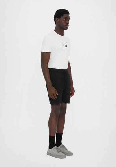 Weißes Baumwoll-T-Shirt mit einem schwarzen Grafikdruck, kombiniert mit schwarzen Shorts. Graue Wildlederschuhe und schwarze Socken runden den Look ab.