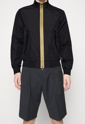 Person, die eine schwarze Jacke mit einem vertikalen beigen Reißverschluss und dunkelgraue plissierte Shorts trägt, vor einem schlichten hellen Hintergrund steht.