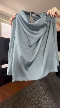 Hände halten eine hellblaue satinierte ärmellose Bluse mit einem drapierten Halsausschnitt und einem Ausschnitt mit Knopfverschluss auf der Rückseite.
