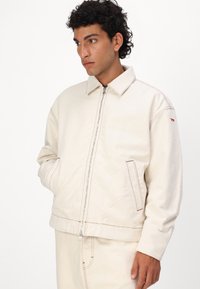 Chaqueta de denim blanca con cierre de cremallera, cuello puntiagudo y detalles de costura roja. Cuenta con bolsillos laterales y un diseño de corte relajado.