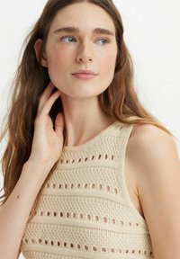 Canotta in maglia beige con un motivo testurizzato che presenta sezioni solide e aperte alternate. Scollo rotondo e design senza maniche.