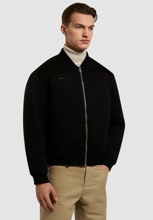Giubbotto bomber nero con zip frontale, polsini e orlo a costine, patch in eco-pelle e texture liscia, indossato sopra un dolcevita bianco.
