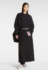 Sweatshirt noir avec logo en relief, jupe noire longue avec textures variées, et baskets rose pâle avec une étiquette à carreaux.