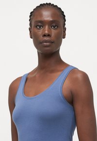 Blå tettsittende tanktopp laget av et glatt, lett stoff. Har en rund hals og smale skulderstropper.