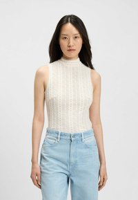 Kobieta z długimi ciemnymi włosami, ubrana w bezrękawnik z białej koronkowej tkaniny w wzory oraz jasnoniebieskie dżinsy z wysokim stanem, stojąca na tle jednolitym.