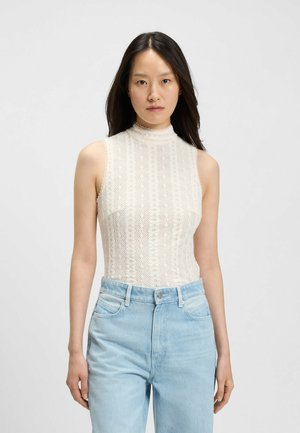 Vrouw met lang zwart haar, gekleed in een mouwloze witte bovenkant met een patroon van kant en lichte blauwe high-waisted jeans, staand tegen een effen achtergrond.