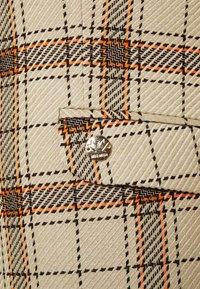 Tissu beige à motif à carreaux noir et orange ; comprend une poche poitrine et un bouton métallique de marque. Surface texturée avec des lignes diagonales.