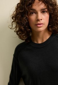 Maglione nero a maglia con collo a girocanale, che presenta una texture morbida e un design aderente. Il modello ha i capelli ricci e un'espressione neutra.