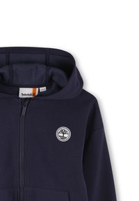 Felpa con cappuccio di colore blu navy, dotata di zip completa, interno morbido e logo alberi Timberland bianco sul petto.