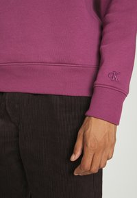 Felpa viola con polsini a coste, caratterizzata da un logo ricamato sulla manica. Abbinata a pantaloni in velluto a coste scuri, che mettono in mostra un tessuto testurizzato.