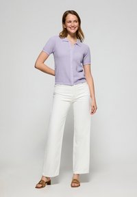 Lila polo met korte mouwen en een gestructureerd patroon, gecombineerd met witte wijde broek en bruine sandalen met straps, staand voor een neutrale achtergrond.