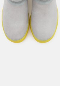 Ett par ljusgrå mockaskor med klara gula gummisulor sett uppifrån mot en vit bakgrund.