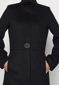 Abrigo negro con un cuello alto, presenta un diseño tonal, mangas largas y un detalle de botón plateado redondo en la cintura.