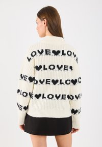 Pletaná kardigan v krémovej farbe s čiernym textom "LOVE" a vzormi srdiečok. Má rebrovaný lem a mierne oversized strih.
