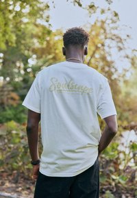 T-shirt en coton blanc avec "Soulscape" en écriture dorée dans le dos, présentant une coupe décontractée et des manches courtes, associé à un short foncé.
