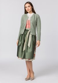 Hellgrüne Strickjacke mit Kabelmuster, pinke Bluse mit Knöpfen, satinierter grüner Rock mit Band, florale Fütterung, beigefarbene Schuhe.