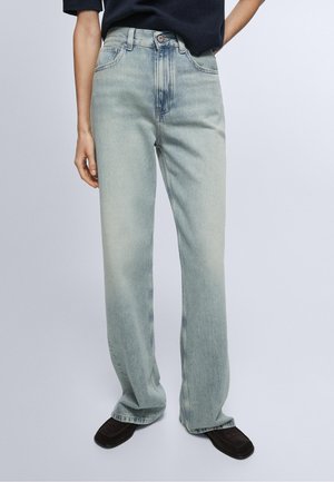 Persoon die wijde lichtblauwe jeans en zwarte schoenen met vierkante neus draagt, staand tegen een effen lichte achtergrond.