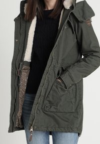 Olivově zelená parka bunda s kapucí podšitou kožešinou, zapínáním na zip a patentky, bočními kapsami a květinovým vzorem uvnitř podšívky.
