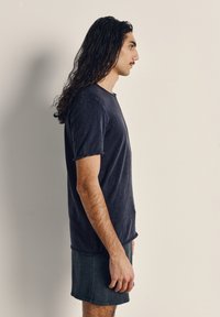 Ung man med långt lockigt hår står i profil vänd mot höger, klädd i en mörk t-shirt och shorts mot en enkel ljus bakgrund.