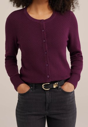 Femme portant un cardigan violet texturé boutonné à manches longues, rentré dans un pantalon noir avec une ceinture noire à boucle dorée.