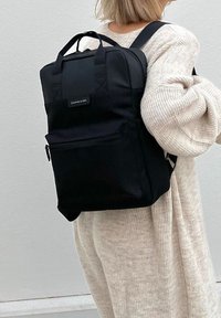 Osoba w beżowym swetrze z dzianiny niesie czarny prostokątny plecak z górnymi uchwytami i przednią kieszenią na zamek.