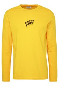 Langärmliges gelbes T-Shirt aus Baumwolle mit einem schwarzen Grafikdruck auf der Brust und einem Rundhalsausschnitt. Glatte Textur.