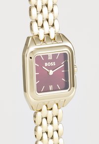 Montre-bracelet dorée avec un boîtier carré, un cadran bordeaux, des index horaires blancs et un bracelet en maillons métalliques. Porte le nom de la marque "BOSS."