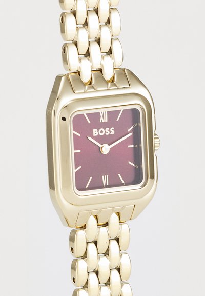 Montre-bracelet dorée avec un boîtier carré, un cadran bordeaux, des index horaires blancs et un bracelet en maillons métalliques. Porte le nom de la marque "BOSS."