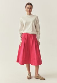 Valge pikkade varrukatega pluus, mida on kombineeritud erksavärvilise roosa volditud midi seelikuga. Modell kannab beeže vabaajajalatsid. Lihtne taust rõhutab rõivastuse detaile.