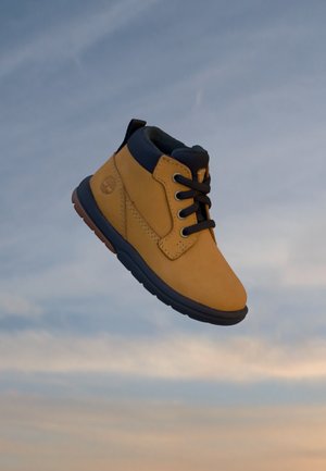 Une seule botte d’enfant couleur fauve et noire avec des lacets noirs flottant sur un fond de ciel bleu et beige.