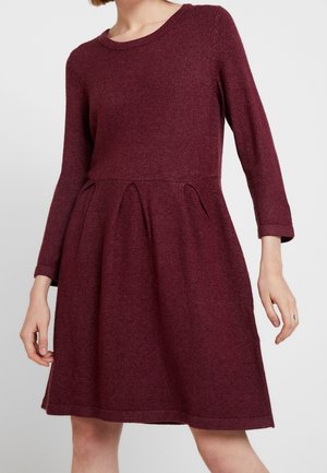 Kobieta ubrana w bordową dzianinową sukienkę z długim rękawem, okrągłym dekoltem i plisowaną spódnicą na jednolitym tle.