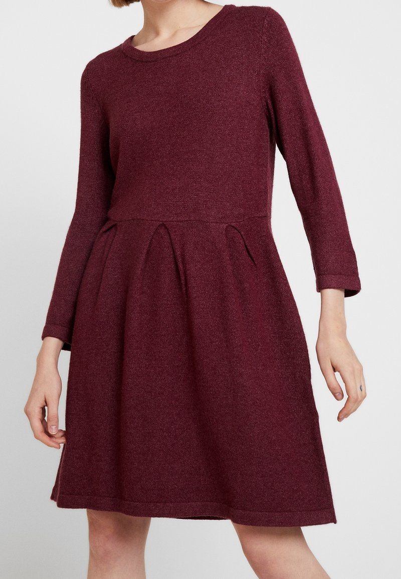Femme portant une robe en maille bordeaux à manches longues avec un col rond et une jupe plissée, sur un fond uni.