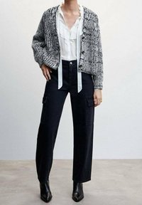 Texturierter grauer und schwarzer Strickcardigan über einer weißen rüschierten Bluse, kombiniert mit hoch taillierten schwarzen Cargo-Hosen und schwarzen Absatzstiefeln.
