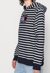 Sudadera a rayas en azul marino y blanco, con un bolsillo en el pecho que presenta acentos en rojo, azul y gris. Puños y dobladillo de canalé, ajuste relajado.
