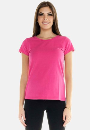 Jeune femme souriante, portant un t-shirt rose vif à manches courtes et un pantalon noir, debout devant un fond clair uni.