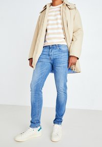 Man i beige huva jacka, beige och vit randig skjorta, ljusblå slim fit jeans och vita sneakers med turkosa detaljer, står på vit golv.