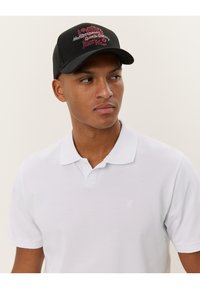 Casquette de baseball noire avec broderie "Mediterranean Sports Series" en rose. Modèle portant un polo blanc uni.