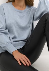Femme portant un pull bleu clair scintillant à manches bouffantes et un legging en cuir synthétique noir, assise avec une jambe pliée.