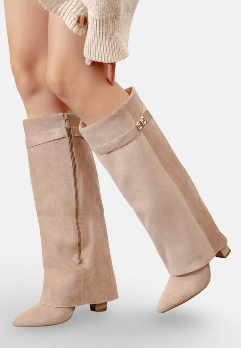 Bottes en suède beige montantes avec un bout effilé, fermeture éclair sur le côté et accents dorés décoratifs au niveau du bord. Texture lisse et légère talon.
