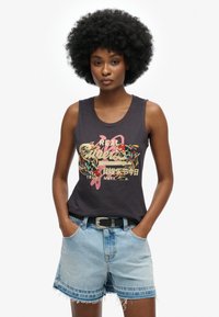 T-shirt sans manches noire avec un design graphique coloré comportant des éléments floraux et textuels. Associé à un short en denim bleu clair.