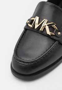 Mocassins en cuir noir avec une boucle logo MK dorée, présentant des détails de couture et une texture lisse. Bout rond et design discret.