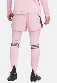 Pantaloni sportivi rosa con un design testurizzato, abbinati a leggins rosa con strisce nere e scarpe da ginnastica nere con dettagli dorati.
