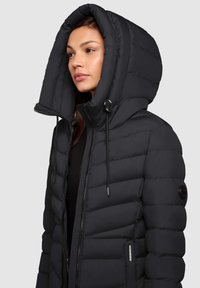 Zwarte pufferjack met een grote, gestructureerde capuchon, zichtbare stiksels en toggles. Voorzien van een rits en een stoffen logopatch op de mouw.