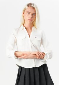 Bijela košulja na dugme s dva džepa na prsima, mekane teksture, zakrivljenim rubom i rolani rukavima koji otkrivaju uzorak pruga. Model je kombinira s plisiranom suknjom.