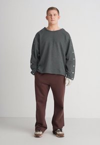 Grå oversized sweatshirt med fremtredende sømmer og knappdetaljer på ermene, kombinert med brune vide bukser og beige sneakers.