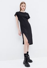 Vestido negro midi con mangas cortas, abertura lateral, textura suave, escote redondo y un sutil detalle del logo. Combinado con botas de combate negras con cordones.